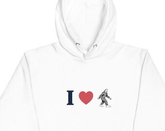 Peut inclure: Sweat-shirt à capuche blanc avec un "I" bleu et un cœur rouge avec une illustration en noir et blanc d'un Bigfoot marchant. Le texte "J'aime Bigfoot" est implicite.