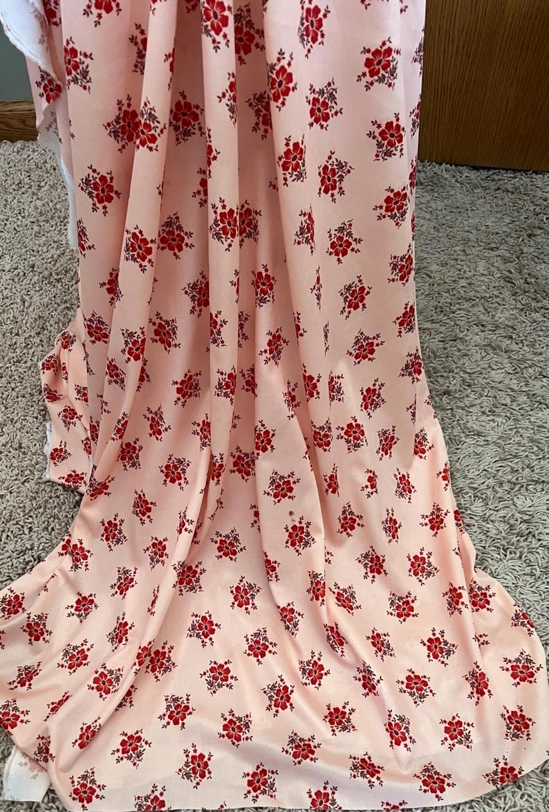 May include: A pink fabric with a repeating pattern of small red flowers. The fabric is folded and draped over a surface.