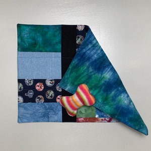 May include: A patchwork dog bed with a blue, green, and black fabric design. A pink and green bone-shaped toy is on top of the bed.