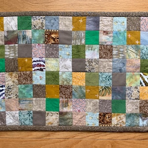 May include: A rectangular patchwork quilt with a brown border. The quilt is composed of numerous small, square fabric patches in various colors, including green, yellow, blue, and gray. The patches feature diverse patterns and textures.