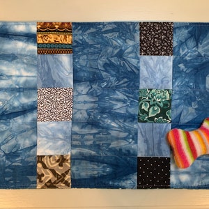 Könnte beinhalten: Ein blaues und weißes Patchwork-Hundebett mit einem gestreiften Knochenspielzeug. Das Bett besteht aus verschiedenen Stoffen, darunter ein blauer Batikstoff, ein weißer Stoff mit schwarzen Punkten und ein grüner Stoff mit Blumenmuster.