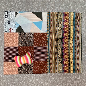 May include: A patchwork quilt with a variety of fabrics and patterns, including brown, tan, blue, and white. A pink and white striped dog toy is in the center of the quilt.