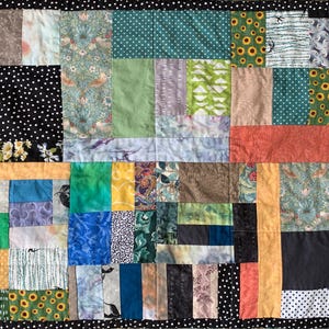 May include: A patchwork quilt with a variety of fabric patterns and colors. The quilt features squares of black polka dots, floral prints, and solid colors like green, orange, and blue. The border is black with white polka dots.