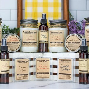 Pode incluir: Uma cole&ccedil;&atilde;o de velas e sprays de ambiente em frascos e garrafas de vidro. As velas s&atilde;o etiquetadas com aromas como Laranja Creamsicle, Flores de Lil&aacute;s, Casa de Praia, P&ecirc;ssego da Ge&oacute;rgia e Coco e Lima. Os sprays de ambiente s&atilde;o etiquetados com aromas como Flores de Lil&aacute;s, Casa de Praia, Ch&aacute; Verde e Erva-Cidreira. Os produtos est&atilde;o dispostos sobre uma superf&iacute;cie branca com um fundo listrado marrom e branco.