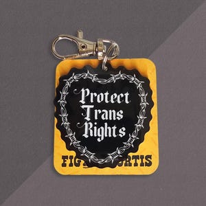 Puede incluir: Un llavero con una base cuadrada amarilla con el texto FIGHT FOR TRANS RIGHTS y un dije de acrílico negro en forma de corazón unido por un broche de metal plateado. El dije negro muestra el texto blanco PROTECT TRANS RIGHTS dentro de un borde de alambre de púas.