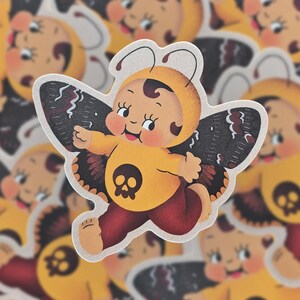 May include: A yellow and black butterfly sticker with a skull design. The butterfly has a human-like face and is wearing a yellow jumpsuit.