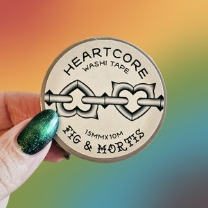 May include: A circular roll of washi tape with a beige label featuring the text HEARTCORE WASHI TAPE and FIG & MORTIS. The label displays a graphic of two heart-shaped links connected by a metal bar and includes the text 15MMX10M.