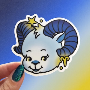 May include: A sticker featuring a cartoon ram with blue horns and a light blue face. The ram has a yellow star above its head and a gold Aries symbol. The sticker is held by a hand with a teal glitter nail.