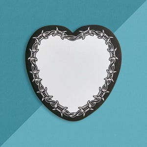 May include: A heart-shaped sticker or paper cutout featuring a solid white center surrounded by a thick black border. The black border is decorated with a detailed white line art illustration of barbed wire that follows the heart shape.