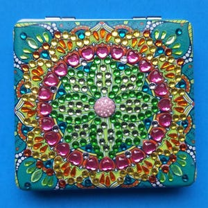 May include: Square compact mirror with a teal background and a colorful, ornate design. The design features a central floral pattern with pink, green, and clear rhinestones, surrounded by orange, yellow, and teal accents. The compact is embellished with a variety of colorful gems.