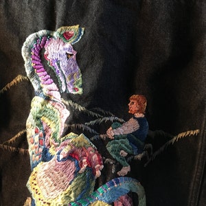 May include: A black denim jacket with a colorful embroidered design of a dragon and a person riding it. The dragon is green, purple, and yellow, and the person is wearing a blue shirt and green pants.