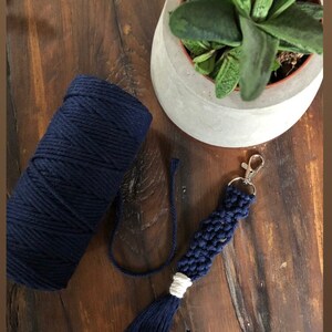 May include: A navy blue macrame keychain with a white tassel and a silver key ring. The keychain is made with cotton cord and is shown next to a ball of navy blue cord.