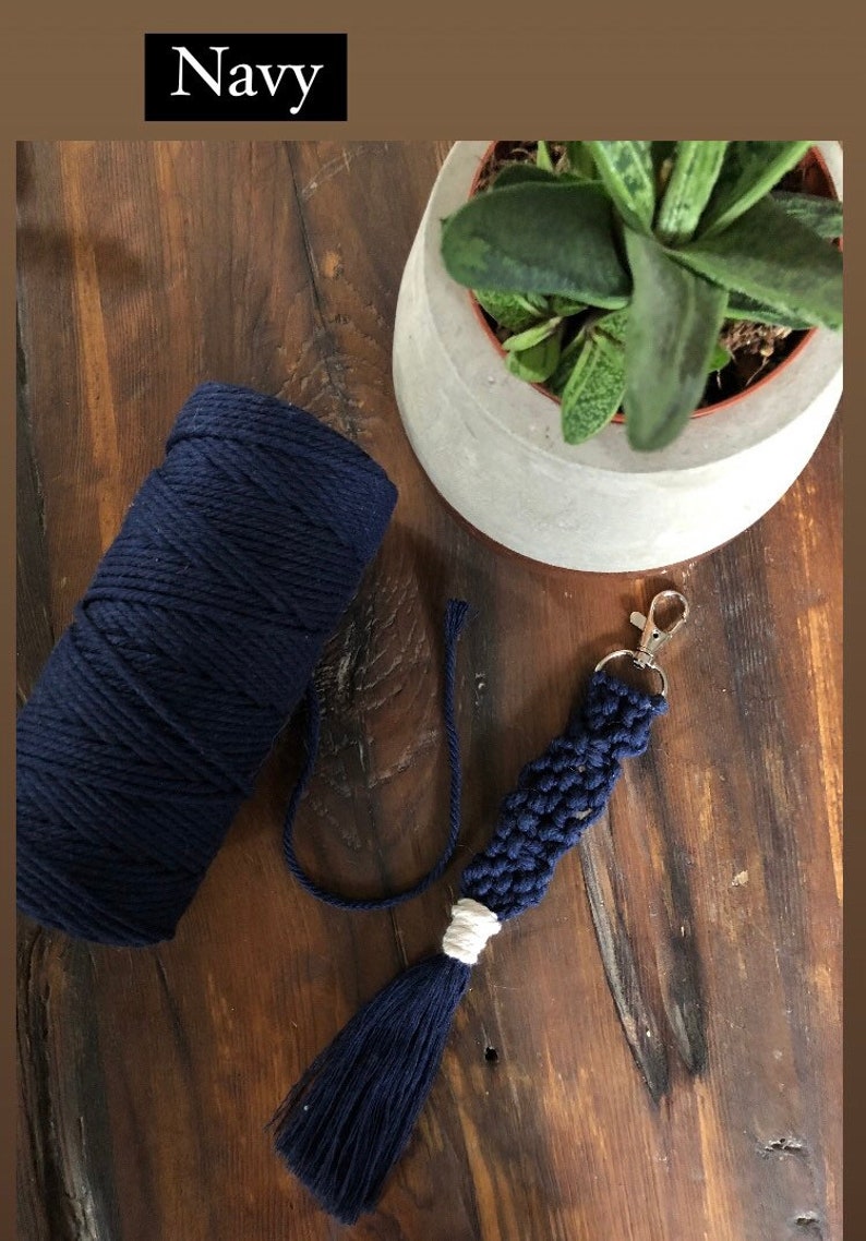 May include: A navy blue macrame keychain with a white tassel and a silver key ring. The keychain is made with cotton cord and is shown next to a ball of navy blue cord.