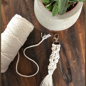 May include: Cream coloured macrame keychain with a tassel and a silver key ring. The keychain is made with a thick cotton cord and is lying on a wooden surface.