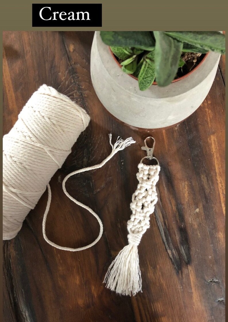 May include: Cream coloured macrame keychain with a tassel and a silver key ring. The keychain is made with a thick cotton cord and is lying on a wooden surface.