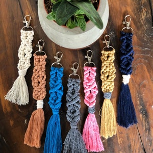 以下が含まれることがあります： タッセルが付いたカラフルなマクラメキーホルダーが7つ。キーホルダーは異なる色のコードで作られており、シルバーのキーリングが付いています。キーホルダーは木製の表面に並べてあります。