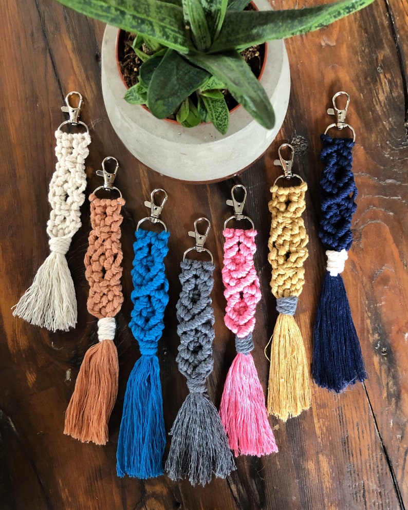 May include: Seven colourful macrame keychains with tassels. The keychains are made with different coloured cords and have silver key rings. The keychains are arranged on a wooden surface.