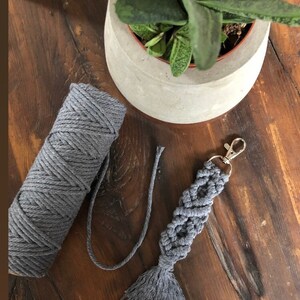 May include: Grey macrame keychain with a tassel and a silver key ring. The keychain is made with a thick grey cord and is shown next to a ball of grey cord.