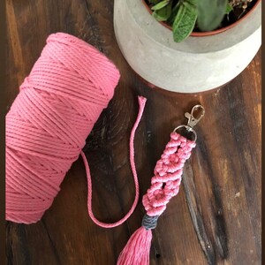 May include: Pink macrame keychain with a tassel. The keychain is made with pink and grey cord and has a silver key ring.