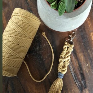 May include: A close-up of a spool of mustard yellow cotton cord with a matching macrame keychain with a blue tassel. The keychain is hanging from a silver key ring.