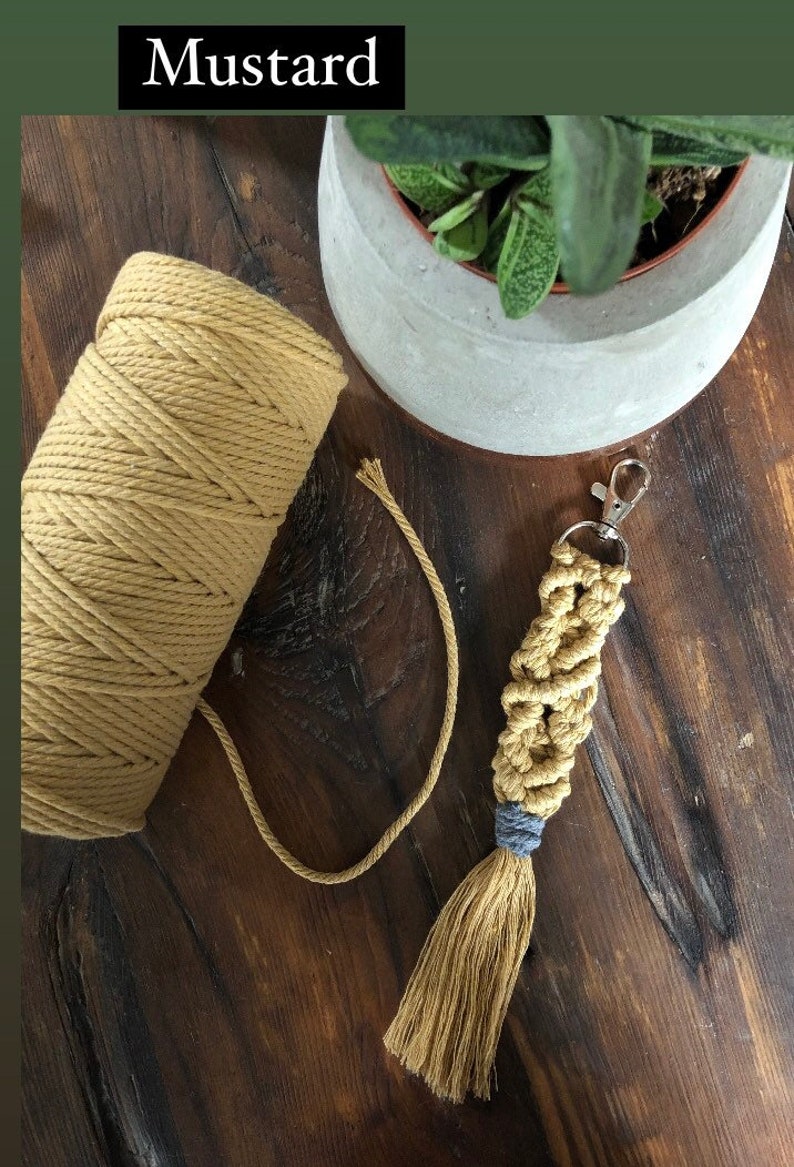 May include: A close-up of a spool of mustard yellow cotton cord with a matching macrame keychain with a blue tassel. The keychain is hanging from a silver key ring.