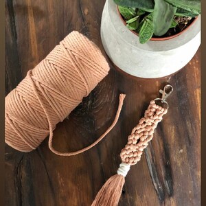 May include: A rust-coloured macrame keychain with a tassel. The keychain is made with a thick cotton cord and has a silver key ring. A ball of the same cord is also pictured.