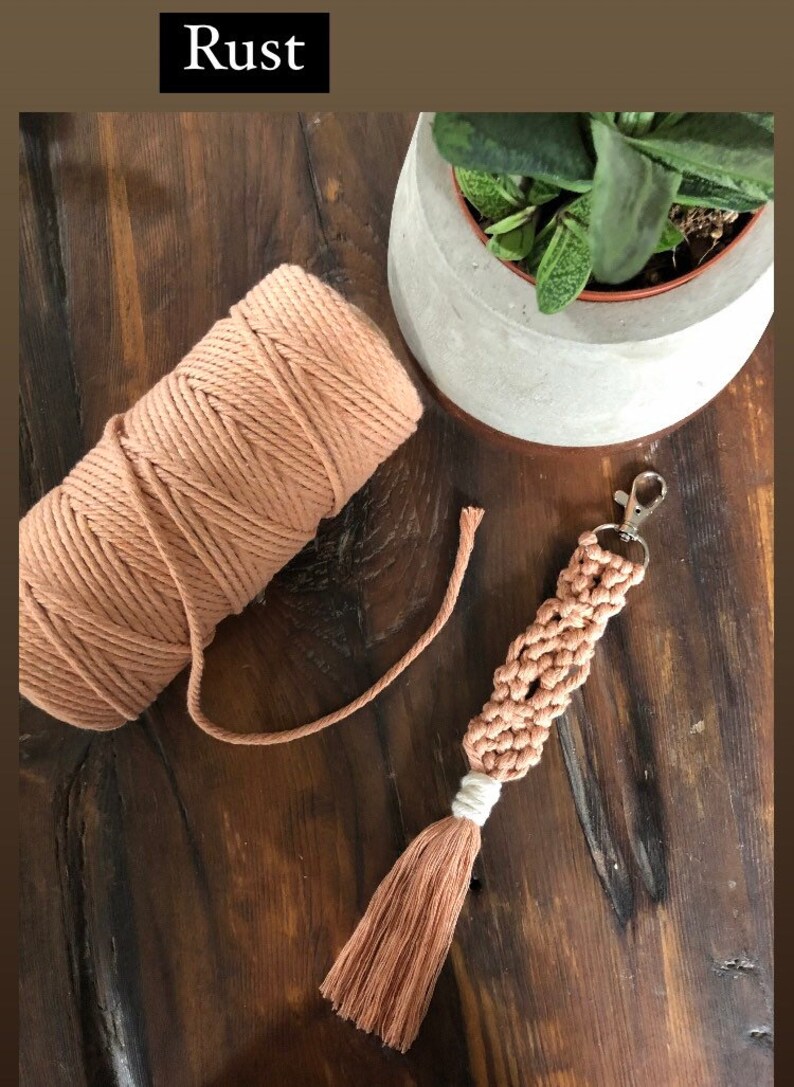 May include: A rust-coloured macrame keychain with a tassel. The keychain is made with a thick cotton cord and has a silver key ring. A ball of the same cord is also pictured.