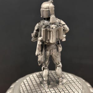 May include: A silver-colored figurine of a character from Star Wars, viewed from the back. The figure is standing on a circular, textured base. The character is wearing a helmet and a jetpack.