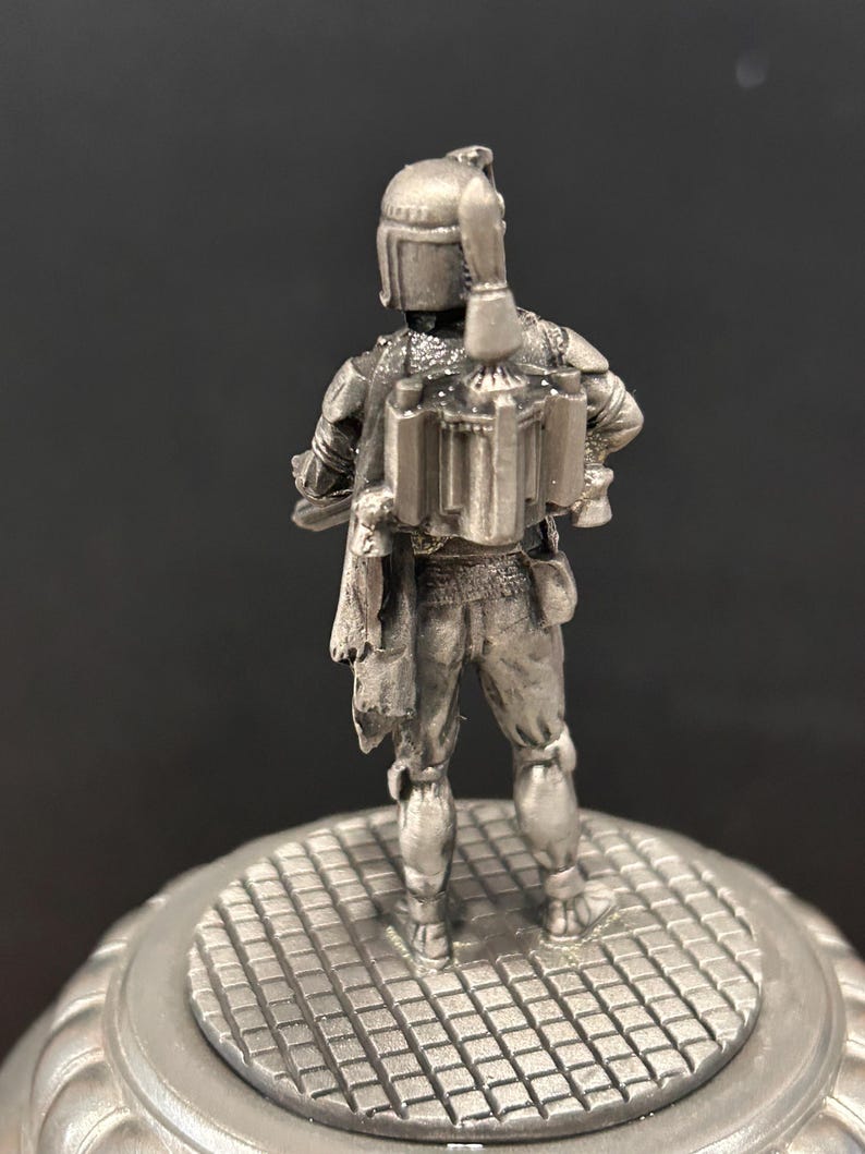 May include: A silver-colored figurine of a character from Star Wars, viewed from the back. The figure is standing on a circular, textured base. The character is wearing a helmet and a jetpack.