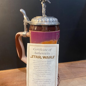 May include: A Star Wars themed beer stein with a pewter figure on the lid. The stein has a brown and tan handle and a certificate of authenticity. The stein is a limited edition item.