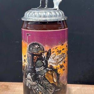 May include: A ceramic beer stein with a detailed illustration of a character from a science fiction film. The stein has a metal lid with a figurine on top. The base of the stein has a decorative design.