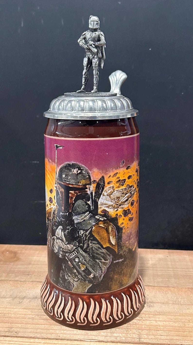May include: A ceramic beer stein with a detailed illustration of a character from a science fiction film. The stein has a metal lid with a figurine on top. The base of the stein has a decorative design.