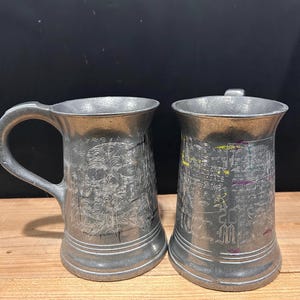 May include: Two silver-toned metal tankards with etched designs. The left tankard features a detailed skull engraving. The right tankard displays text including the year 2018 and various decorative markings with small yellow and purple accents.