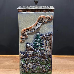 May include: A rectangular ceramic stein with a detailed relief of a castle scene, featuring a banner that reads "Burg-Grenzau." The stein has a metal lid and a dark handle. The color palette includes blues, greens, browns, and creams, creating a vintage aesthetic.