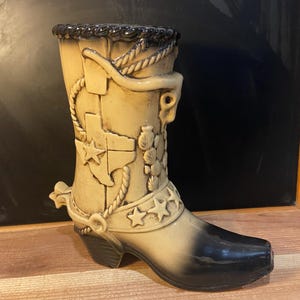 May include: A ceramic cowboy boot vase with a beige and black glaze. The boot features a raised design of a rope, stars, and the state of Texas.