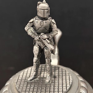 May include: A detailed, silver-colored figurine of a character from a science fiction franchise, standing on a textured, circular base. The figure is clad in armor and holds a weapon, with a helmet obscuring the face. The background is dark.