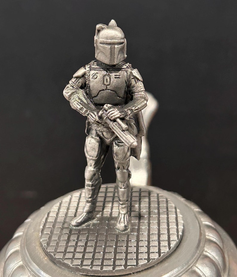 May include: A detailed, silver-colored figurine of a character from a science fiction franchise, standing on a textured, circular base. The figure is clad in armor and holds a weapon, with a helmet obscuring the face. The background is dark.