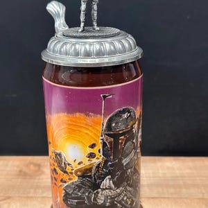 May include: A cylindrical beer stein with a detailed metal lid and handle. The body features a colorful graphic of a character in a helmet against a sunset backdrop. A small metal figurine of the character stands atop the lid.