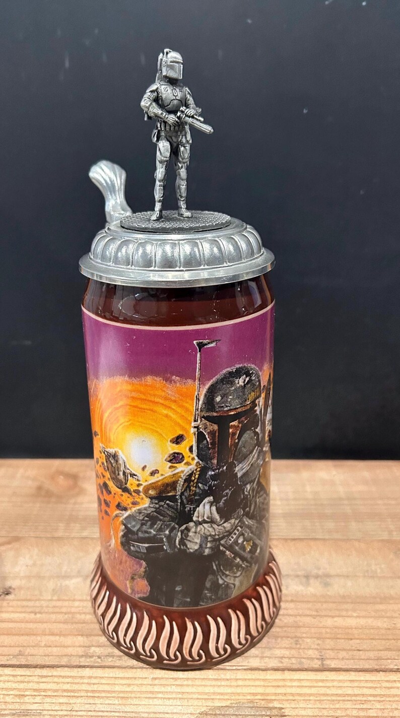 May include: A cylindrical beer stein with a detailed metal lid and handle. The body features a colorful graphic of a character in a helmet against a sunset backdrop. A small metal figurine of the character stands atop the lid.