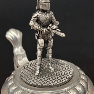 May include: A pewter-colored figurine of a character from a science fiction film, standing on a decorative beer stein lid. The figure is holding a blaster and is wearing a helmet and armor. The stein has a detailed, textured design.