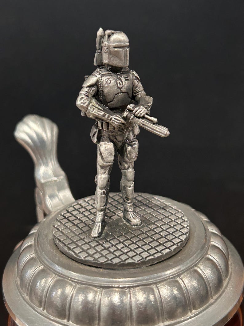 May include: A pewter-colored figurine of a character from a science fiction film, standing on a decorative beer stein lid. The figure is holding a blaster and is wearing a helmet and armor. The stein has a detailed, textured design.