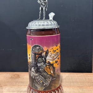May include: A ceramic beer stein featuring a Star Wars theme. The stein has a detailed illustration of a character on the front, a metal lid, and a small figurine on top. The base is brown with a decorative pattern.