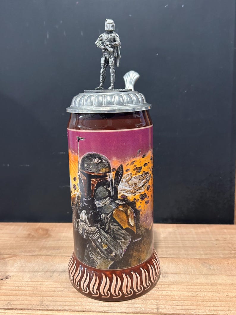 May include: A ceramic beer stein featuring a Star Wars theme. The stein has a detailed illustration of a character on the front, a metal lid, and a small figurine on top. The base is brown with a decorative pattern.