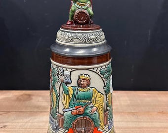 May include: A ceramic beer stein featuring a relief-molded design of a seated king holding a mug while resting on a barrel. The lid is topped with a smaller figurine of a person sitting on a barrel. The stein has brown, green, and gold accents.