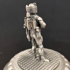 May include: A silver-colored figurine of a character from Star Wars, standing on a round, patterned base. The figure is detailed with a helmet, jetpack, and armor, and is posed with arms crossed. The base has a decorative, ridged edge.