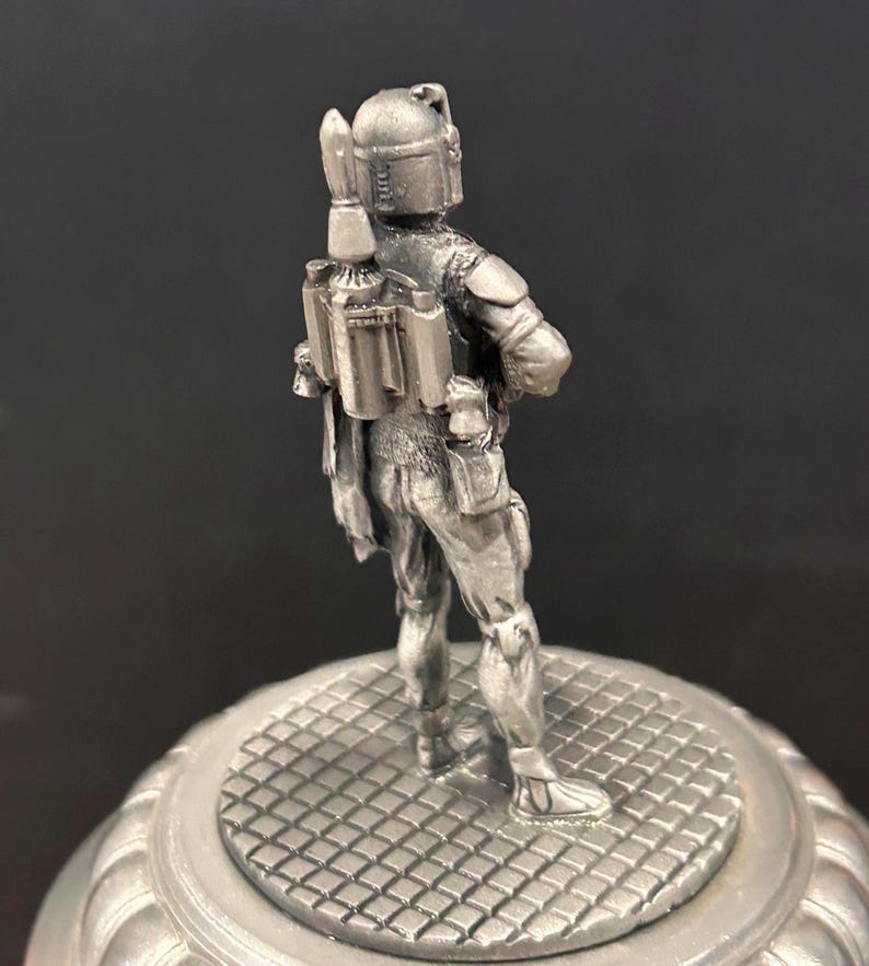 May include: A silver-colored figurine of a character from Star Wars, standing on a round, patterned base. The figure is detailed with a helmet, jetpack, and armor, and is posed with arms crossed. The base has a decorative, ridged edge.