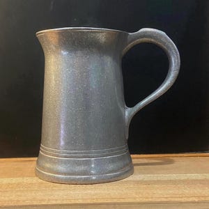 May include: A pewter tankard with a handle, featuring a cylindrical body and a flared base. The tankard has a slightly textured surface and a curved handle for easy gripping. The overall design is simple and classic, suitable for serving beverages.