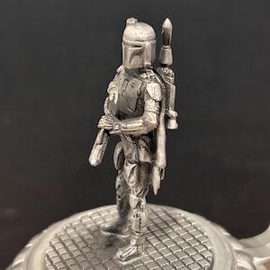 May include: A silver-colored figurine of a character from Star Wars, standing on a textured, circular base. The figure is wearing a helmet and armor, holding a weapon. A backpack is attached to the figure's back. The base has a grid-like pattern.