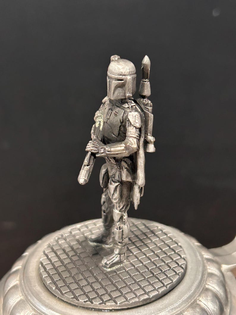 May include: A silver-colored figurine of a character from Star Wars, standing on a textured, circular base. The figure is wearing a helmet and armor, holding a weapon. A backpack is attached to the figure's back. The base has a grid-like pattern.