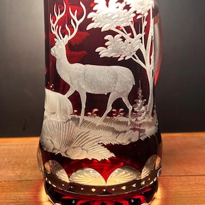 May include: A ruby red glass beer stein with a detailed etched design. The design features a deer in a forest scene with trees and foliage. The stein has a decorative base and a handle.